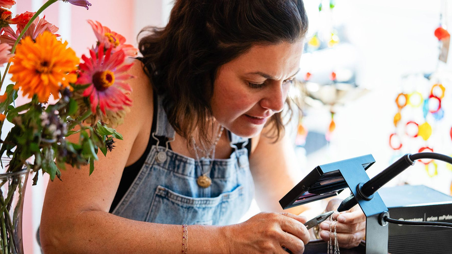 Brandy at Bella Vita Jewelry crafting a permanent jewelry piece in Little Rock, with vibrant flowers in the foreground
