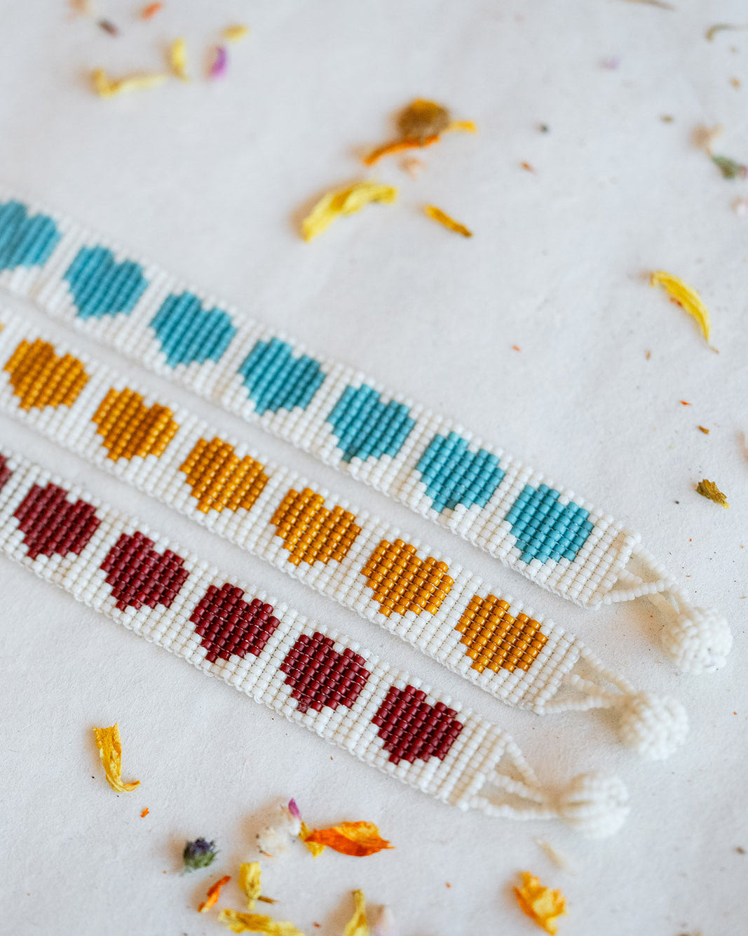 Three handmade beaded friendship bracelets with repeating heart patterns in teal, orange, and burgundy on a white background, styled with dried flower petals.