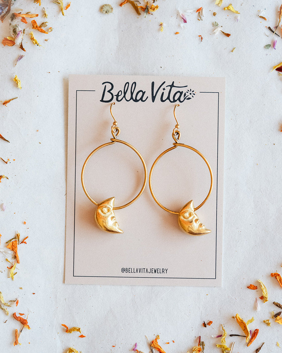 Gold-tone hoop earrings with crescent moon charms, displayed on a Bella Vita jewelry card with dried flower petals on a light surface.