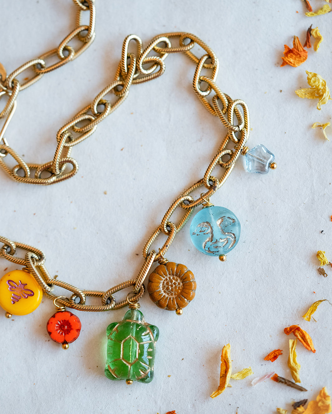 A gold-tone chain necklace with multiple colorful charms, including a green crystal pendant, a blue circular charm, a brown flower charm, and small orange and yellow floral accents, displayed on a white surface with scattered dried flower petals.