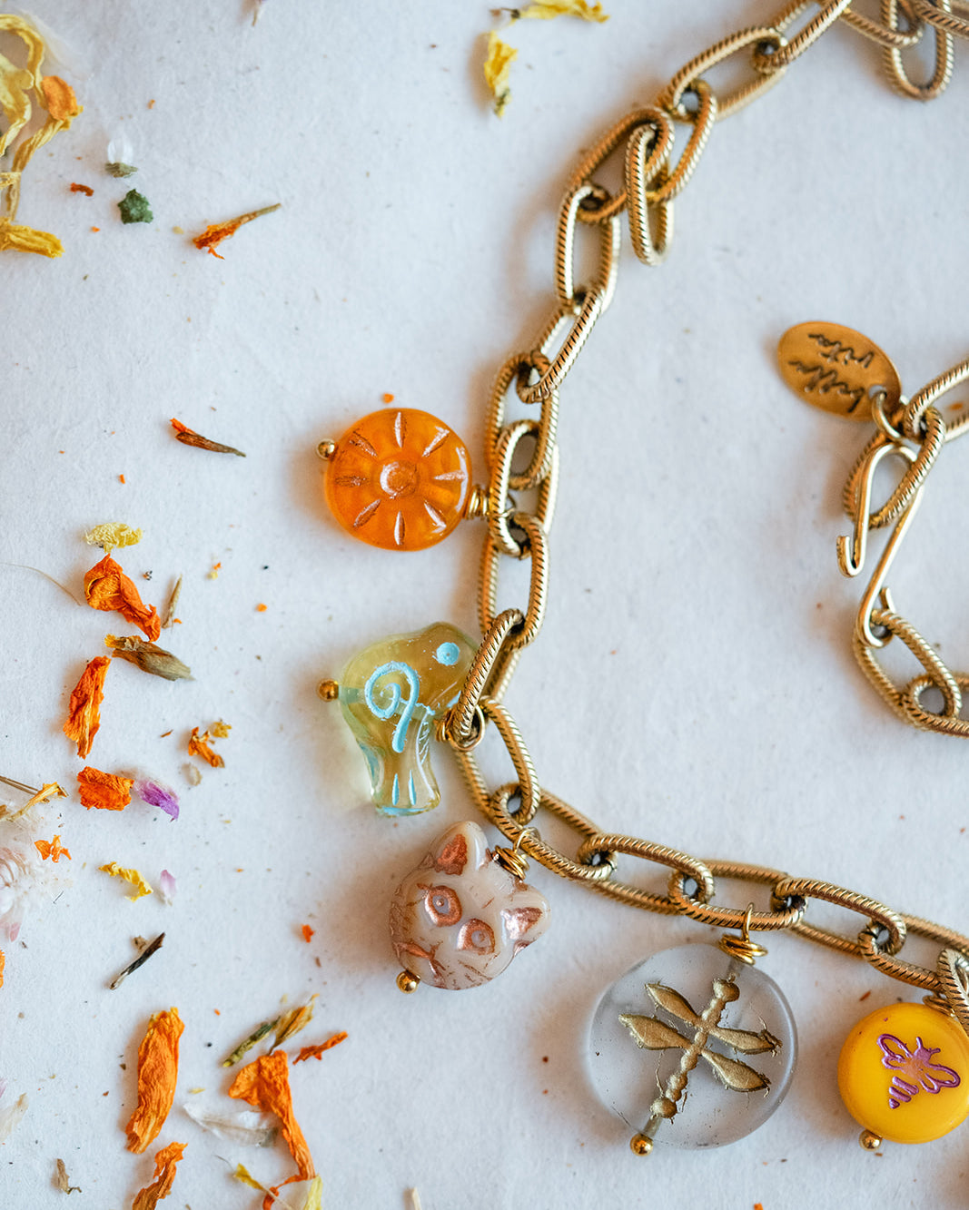 A gold-toned chain necklace with multiple colorful charms, including a sun, a bird, a cat, a dragonfly, and a yellow flower, laid out on a white surface with scattered dried flowers.