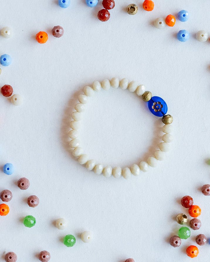 A beaded bracelet made of small off-white beads with a single blue button closure, arranged in a circle on a white background with scattered colorful beads around it.