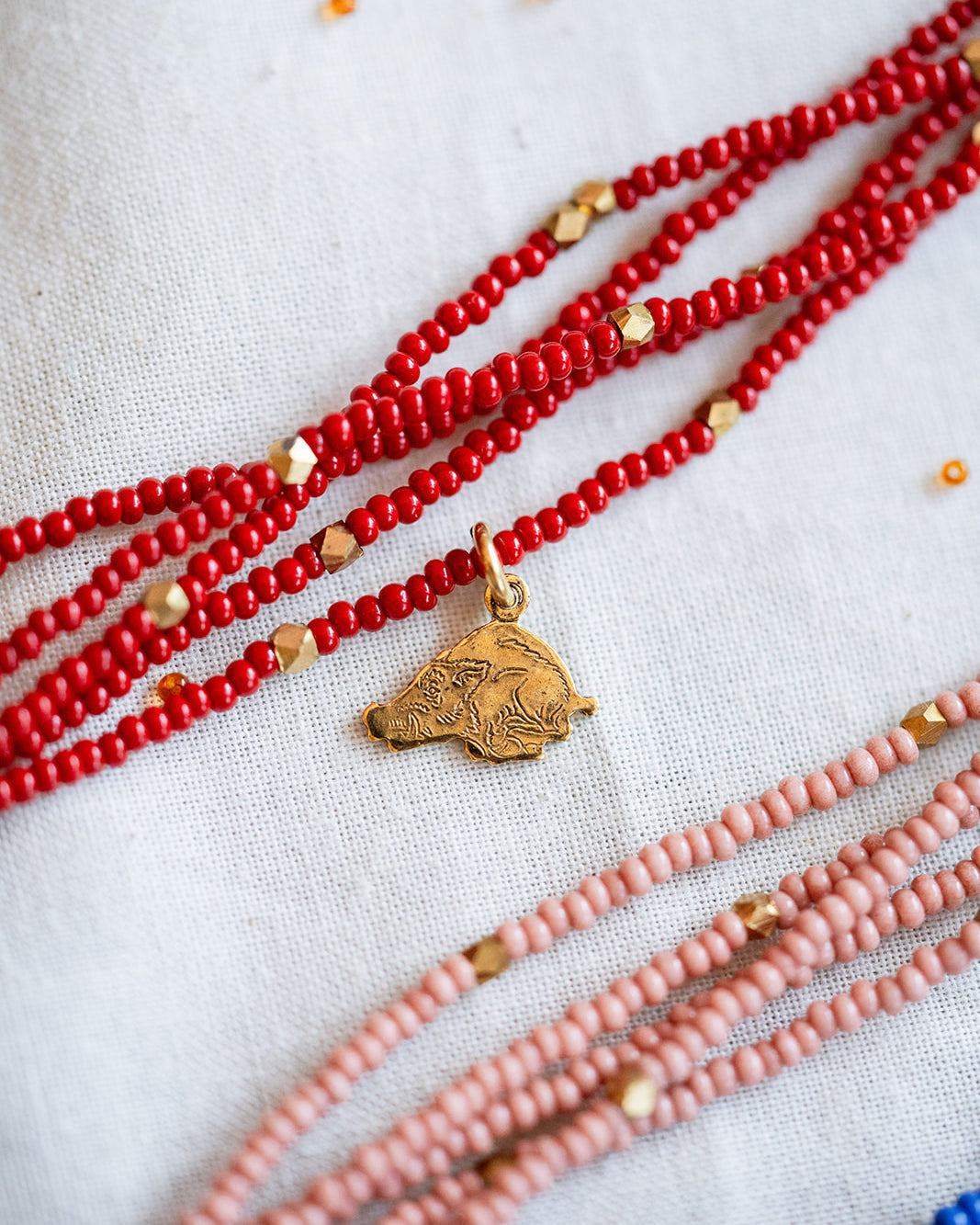 A close-up of multiple beaded necklaces laid out on a light fabric background. The most prominent necklace is made of bright red beads with small gold spacer beads and a small gold pendant.
