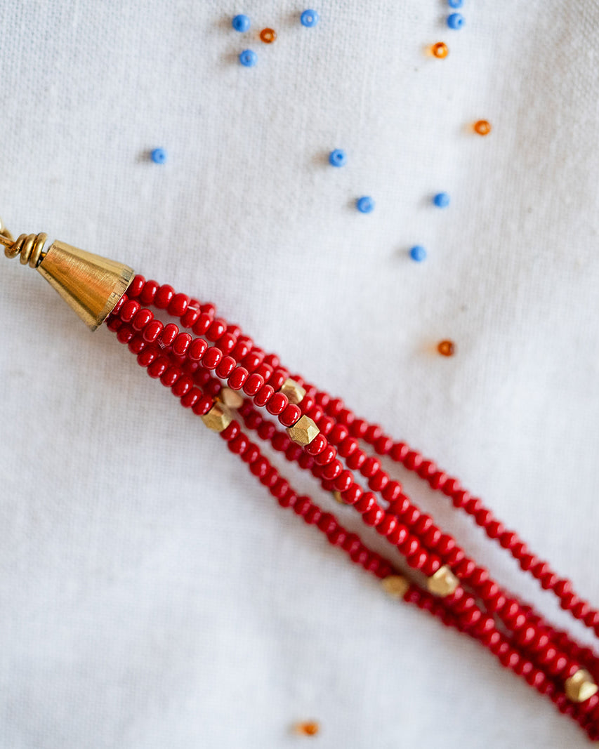 A close-up of a beaded tassel or strap end with multiple strands of red seed beads, a gold cone-shaped connector, and a gold lobster clasp, placed on a white fabric background with scattered small blue and orange beads.
