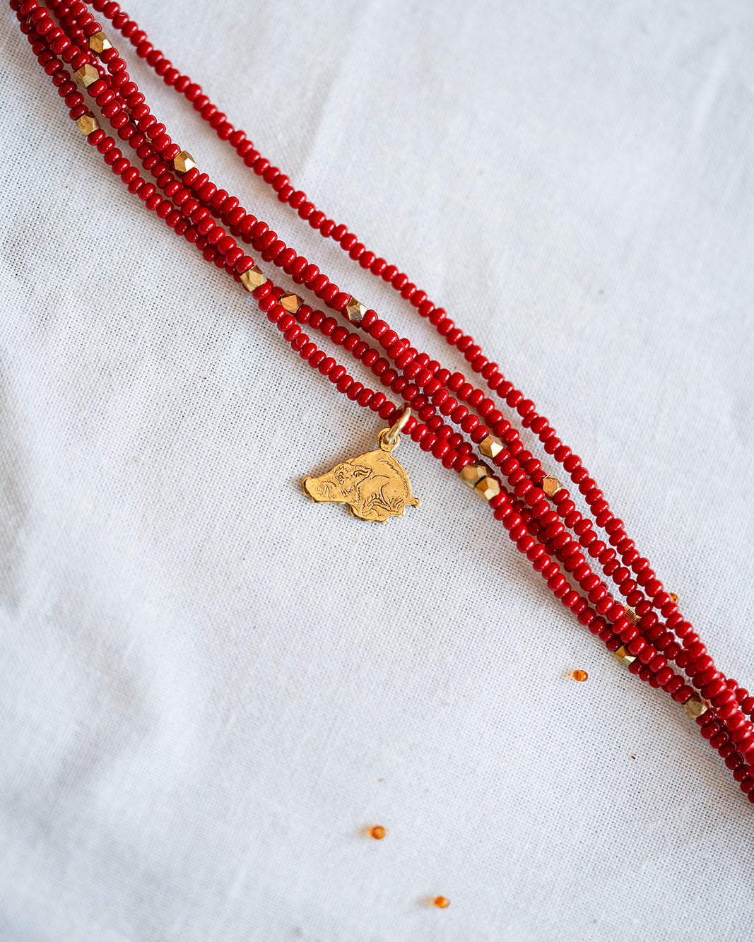 A multi-strand red beaded necklace with small gold spacer beads and a gold pendant, laid diagonally on a white textured fabric background.