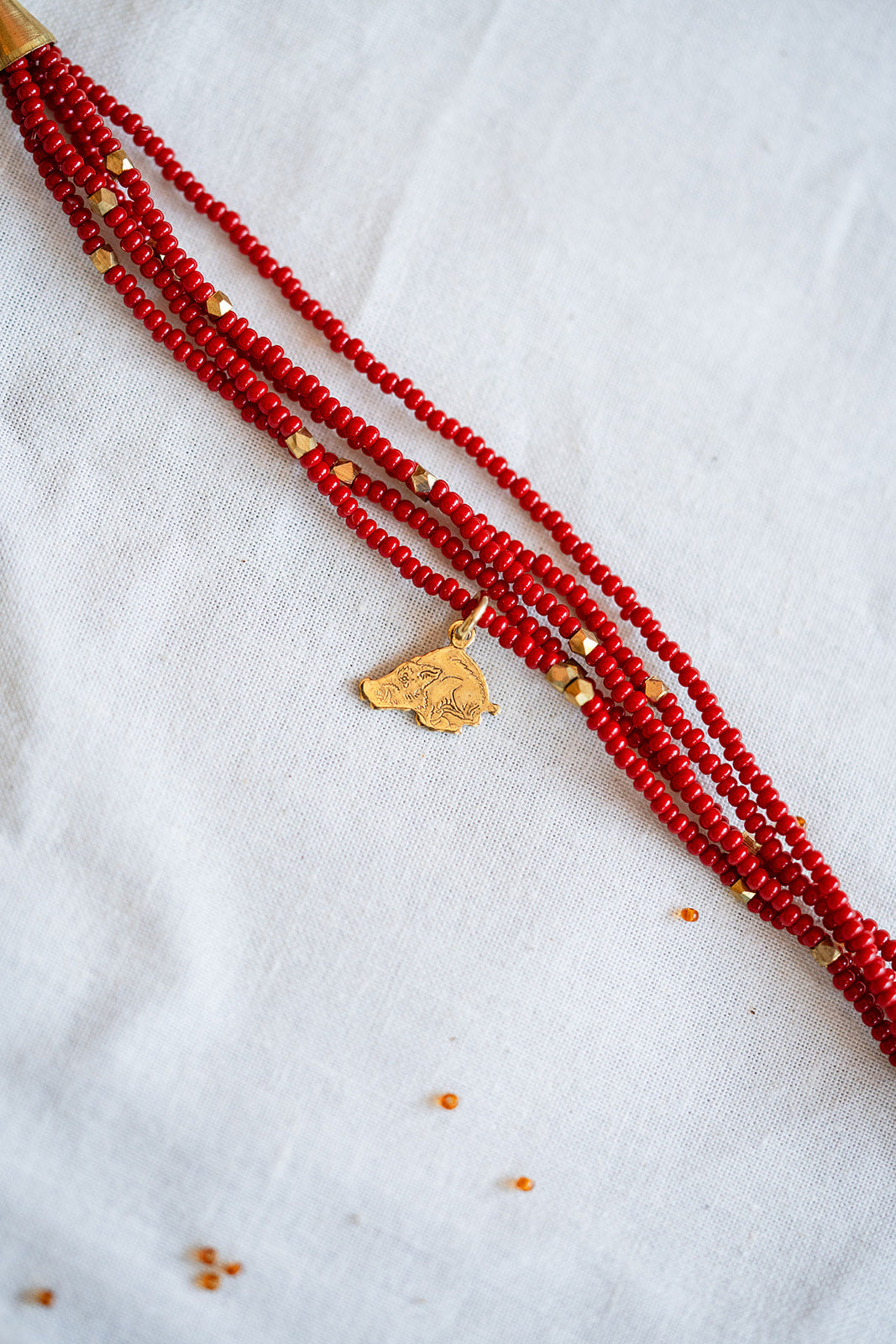 A multi-strand red beaded necklace with small gold spacer beads and a gold pendant, laid diagonally on a white textured fabric background.