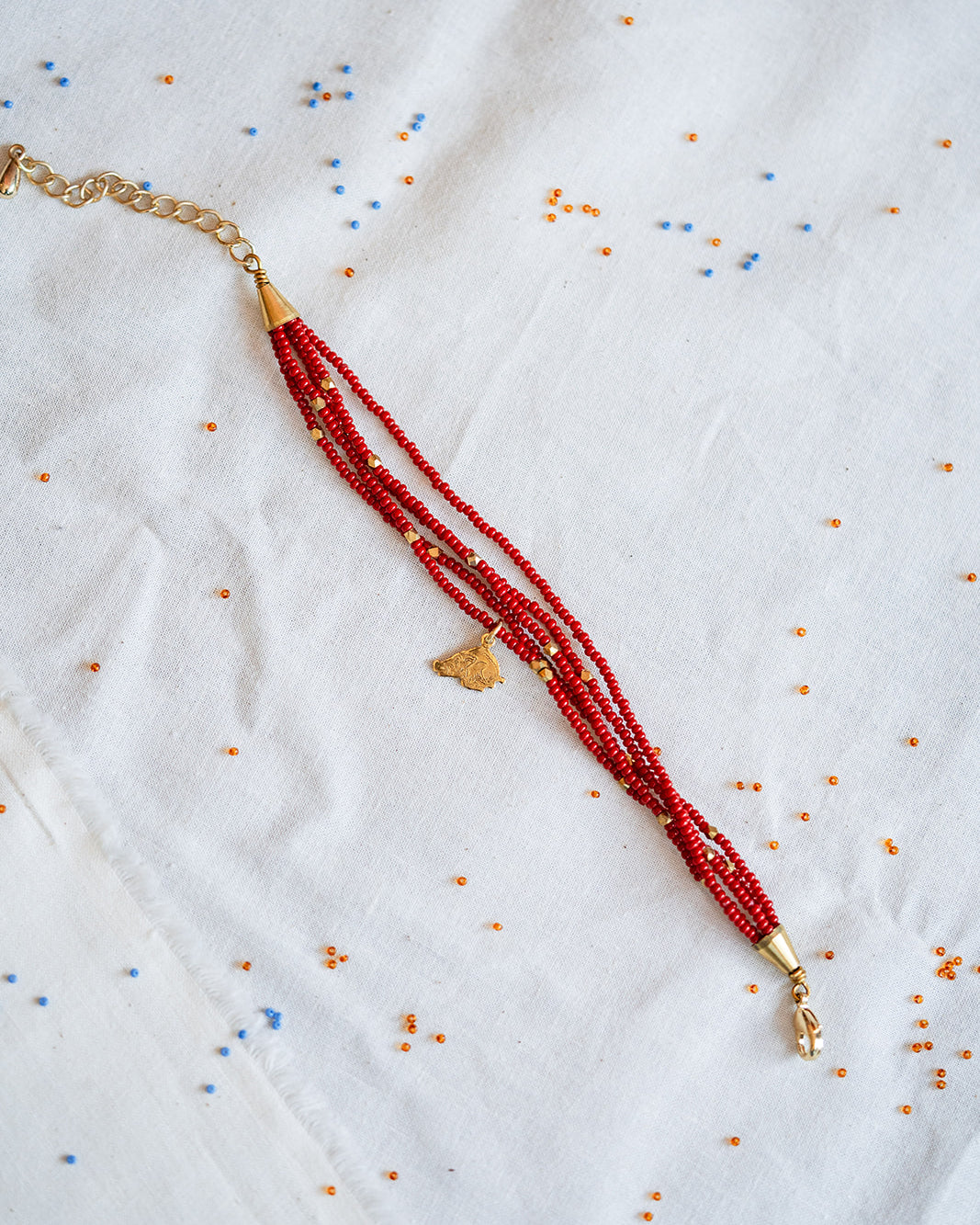 A beaded necklace with multiple strands of red beads, gold spacer beads, and a small gold pendant, laid flat on a white embroidered fabric background.