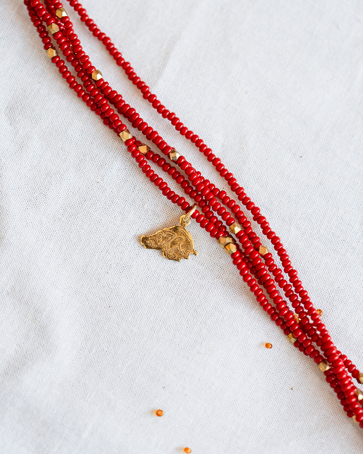 A multi-strand red beaded necklace with small gold spacer beads and a gold-toned pendant, laid diagonally on a white textured fabric background.