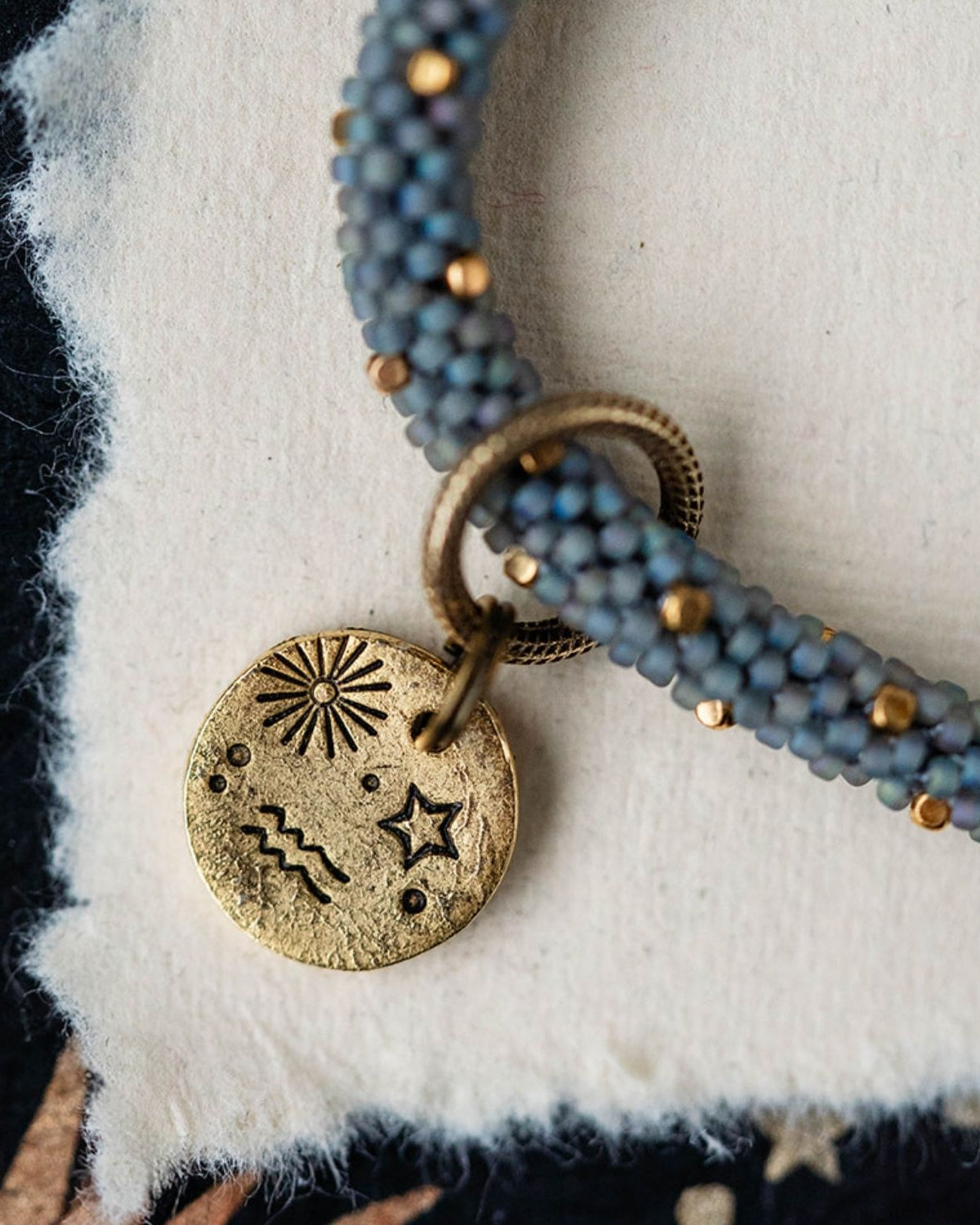 Gold Aquarius pendant attached to blue-grey beaded jewelry strands with gold accent beads detail view
