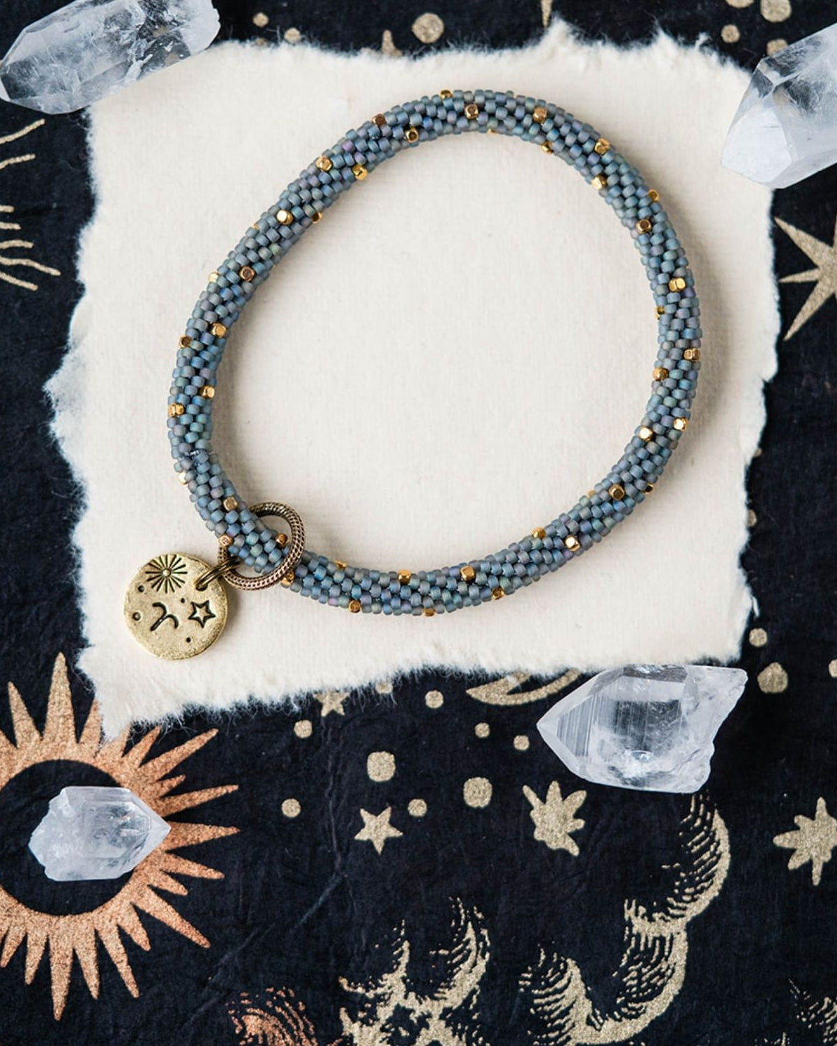 Blue-grey beaded bracelet with gold spacers and Aries zodiac charm on starry background
