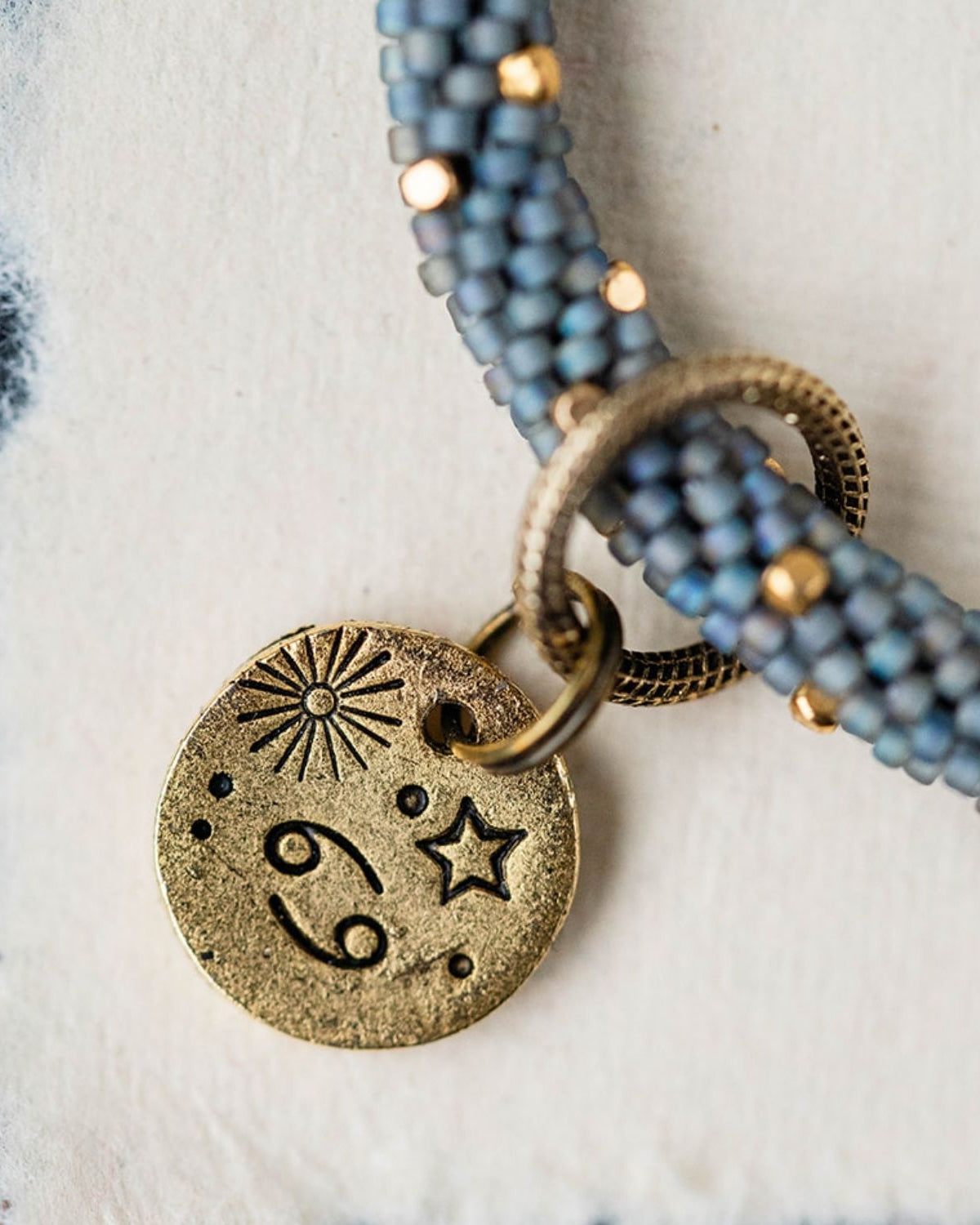 Gold Cancer charm with blue-grey beaded jewelry showing crab constellation and sun engravings