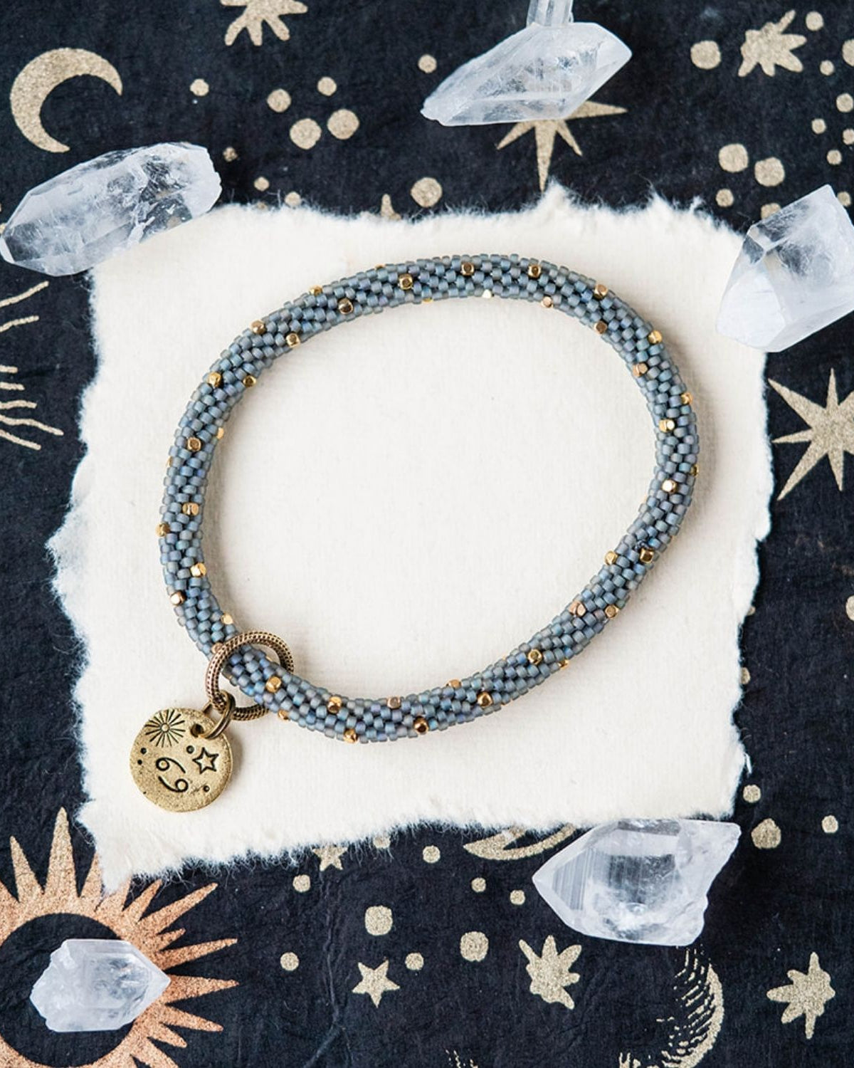 Blue-grey beaded bracelet with gold accents and Cancer zodiac charm on celestial background