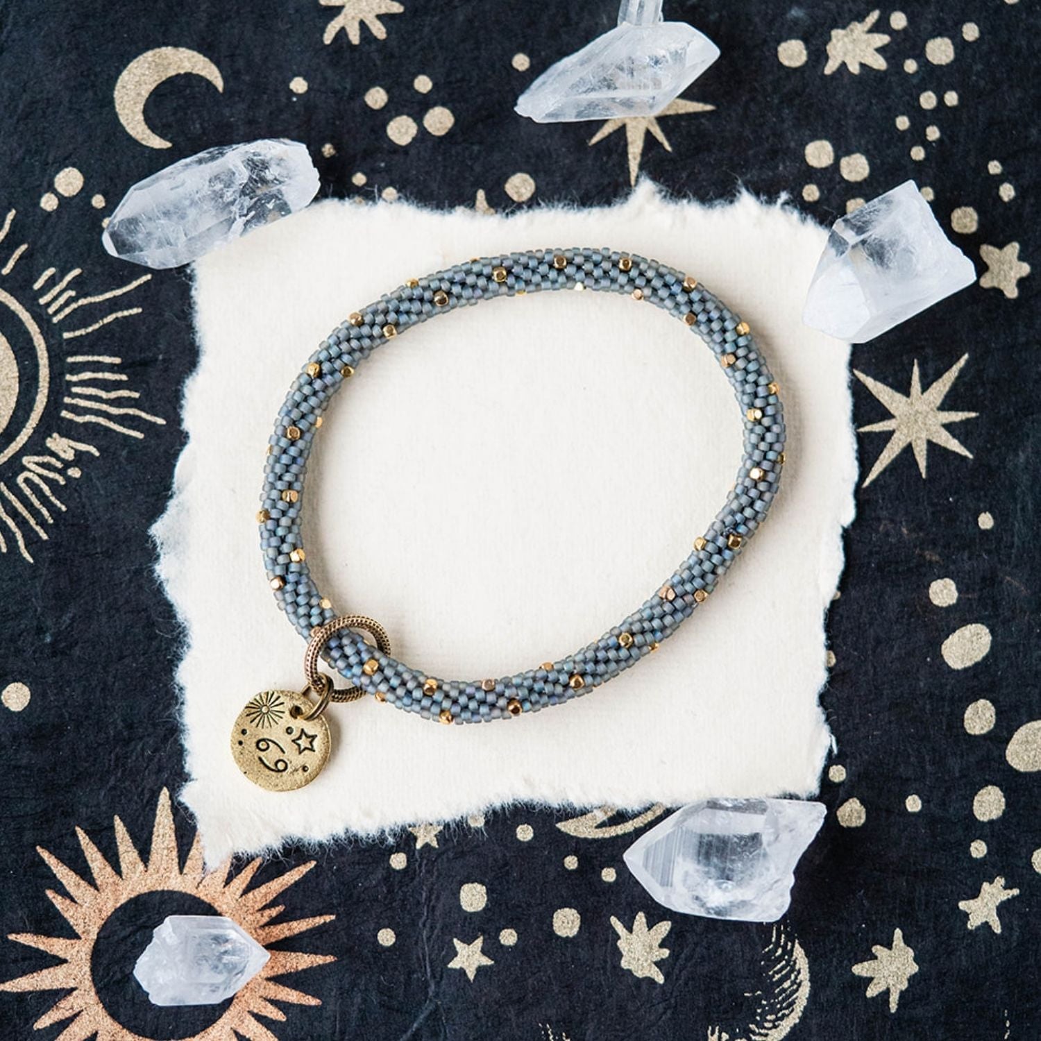 Blue-grey beaded bracelet with gold accents and Cancer zodiac charm on celestial background