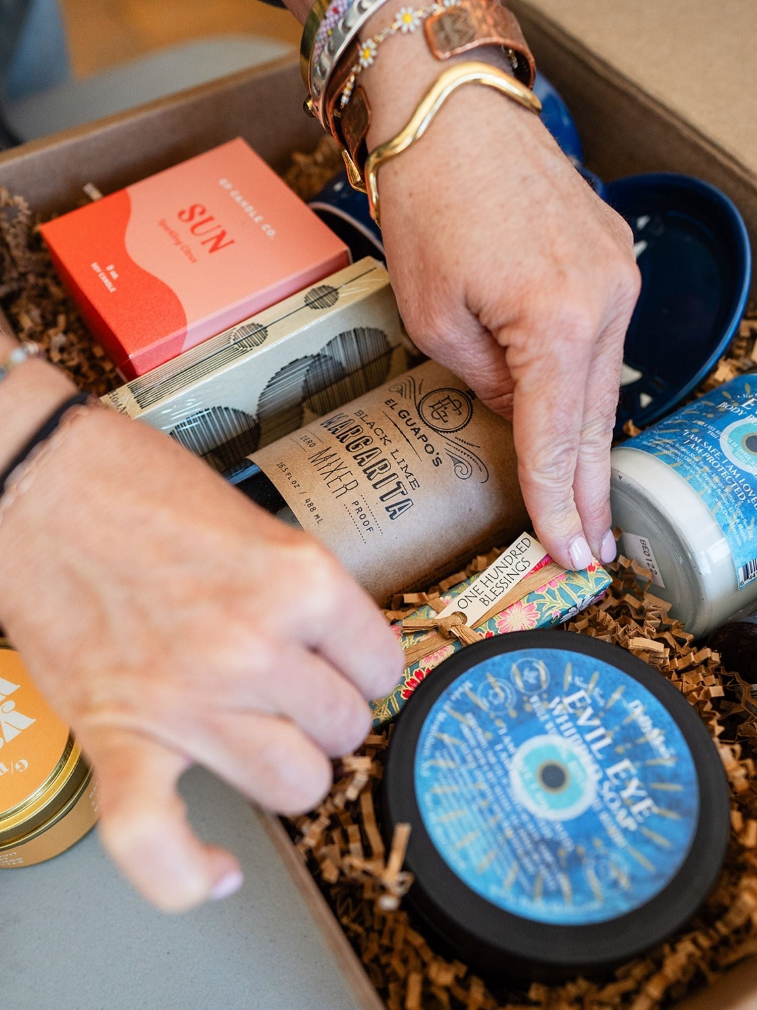 Hands with handcrafted Bella Vita bracelets arranging wellness products in a custom gift box, showcasing jewelry alongside self-care items.