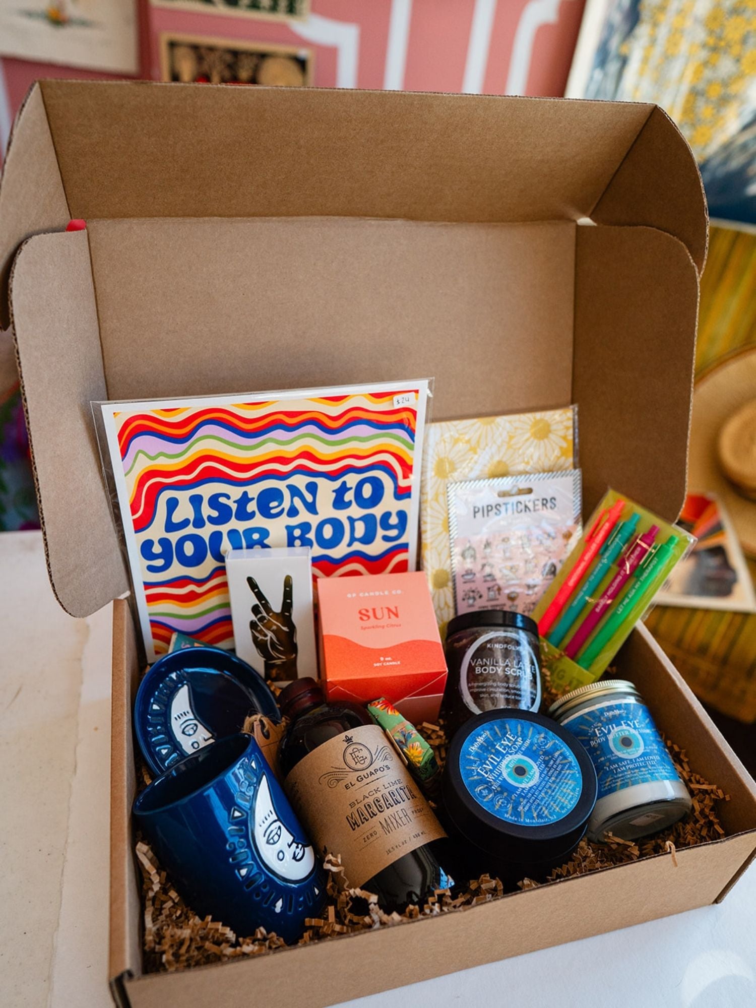 Bella Vita custom self-care gift box featuring Evil Eye body products, blue face mugs, motivational art, and handcrafted items nestled in crinkle paper.