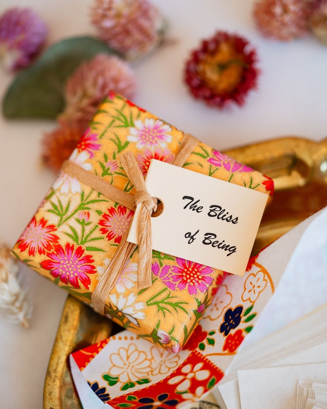 Close-up of 'The Bliss of Being' book with vibrant floral cover and attached tag on gold surface.