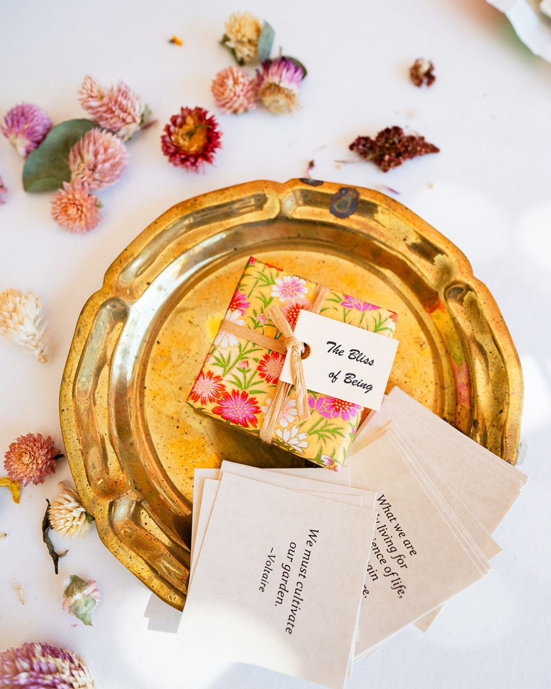 Floral-covered 'The Bliss of Being' book with info cards on gold tray, surrounded by dried flowers.