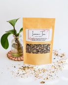 BWG Immuni-tea loose leaf blend in kraft packet with plant in glass jar and scattered tea leaves.