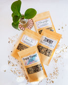 Assorted BWG tea packets with small plant in glass jar on beaded coaster with loose tea sprinkled around.