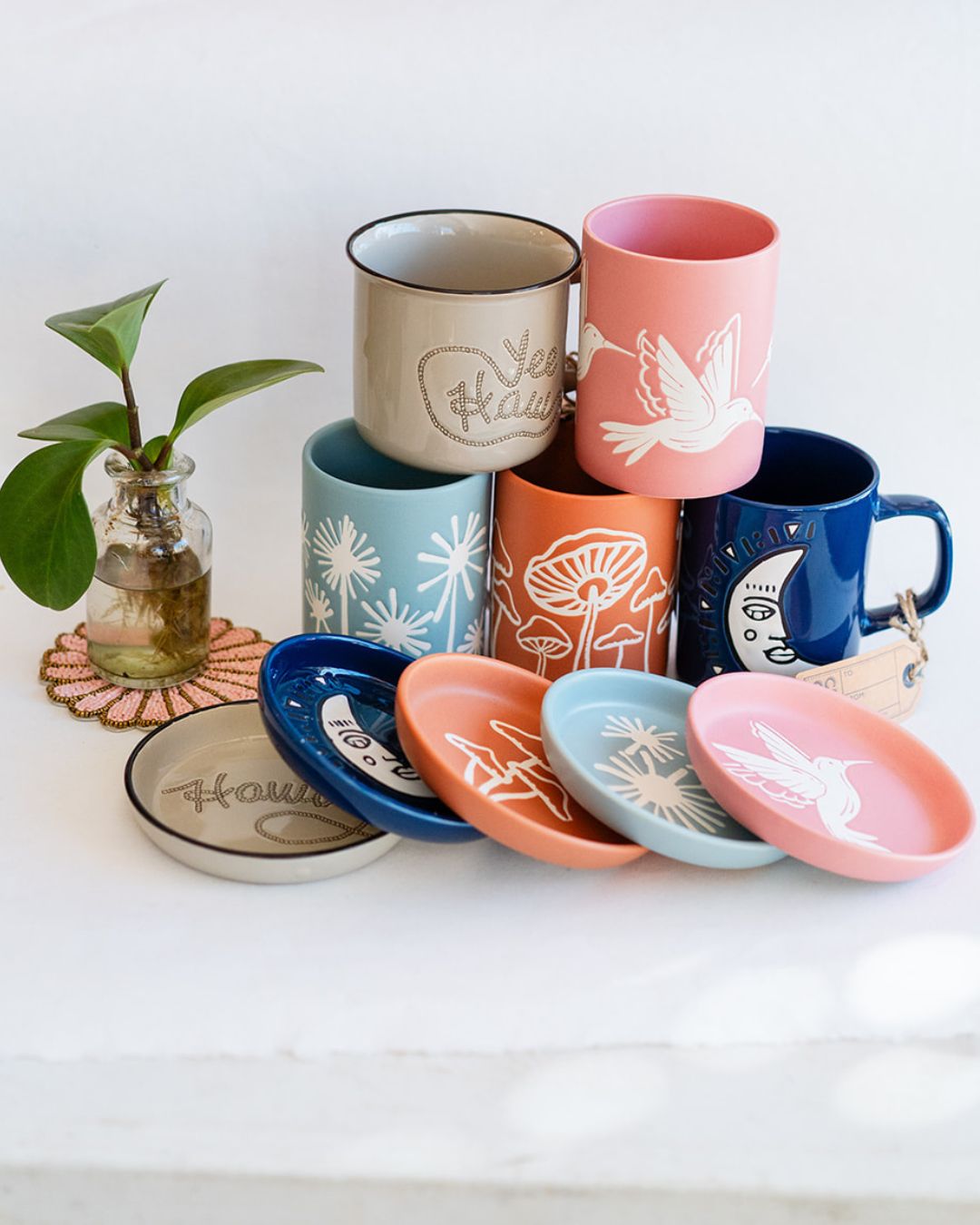 Collection of Cuppa Color ceramic mugs with matching coasters and potted plant