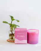 Flora 9oz Hue Candle in pink with Wild Orchid scent, shown with matching box and potted plant on white surface.