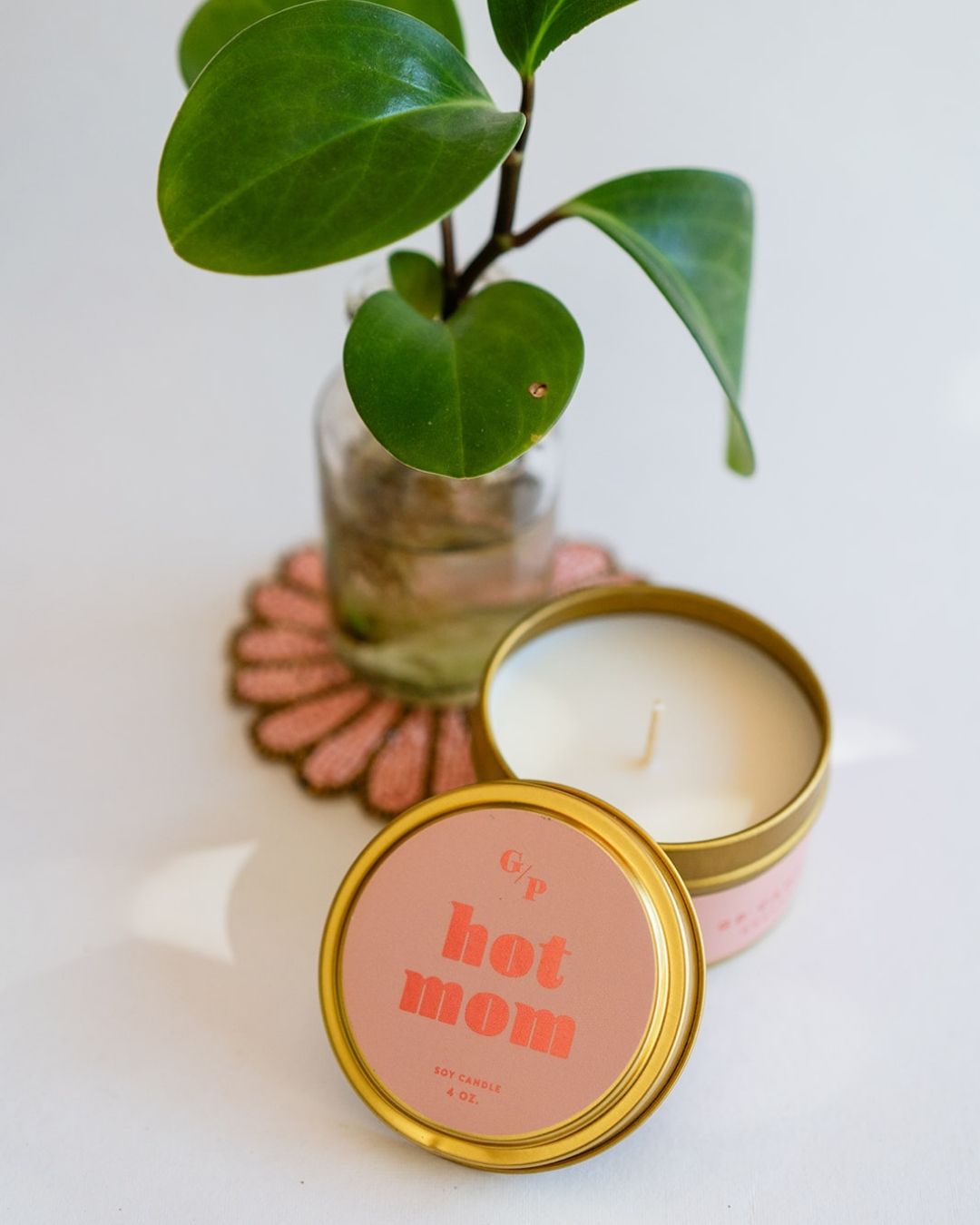 Hot Mom Just Because 4oz candle tin with pink and coral label, displayed alongside small potted plant on decorative coaster.