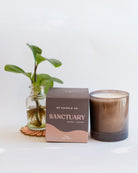 Sanctuary 9oz Hue Candle in brown with Leather & Incense scent, displayed with packaging and small potted plant.