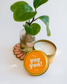 Yay You! Just Because 4oz candle tin with bright orange and white text, styled with small plant on a pink beaded coaster.