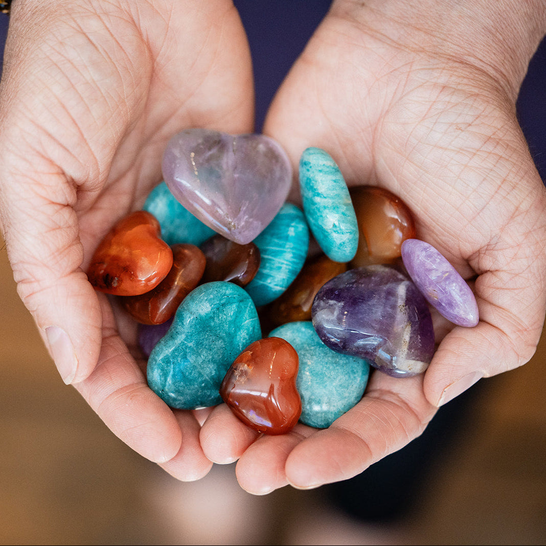 Hands holding assorted semiprecious gemstone pocket hearts including amethyst, turquoise, red jasper, and clear quartz in various polished finishes.