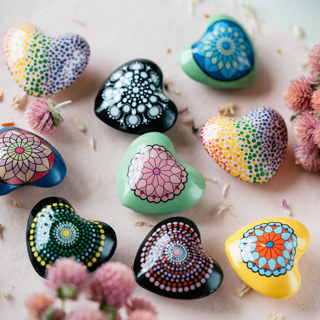 Handpainted mandala jingle hearts in multiple colors featuring intricate dot patterns and floral designs, arranged with dried flowers on soft background.
