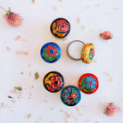 Flatlay collection of Song of India decorative brass solid perfume containers showing varied artistic designs with floral motifs in black, blue, yellow and red, styled with dried flowers.
