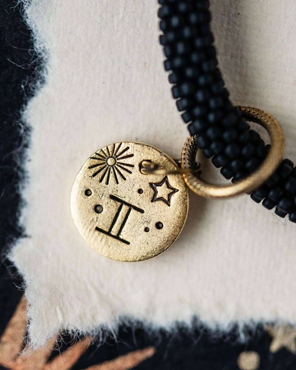 Close-up gold Gemini zodiac charm with twins symbol attached to black beaded jewelry detail