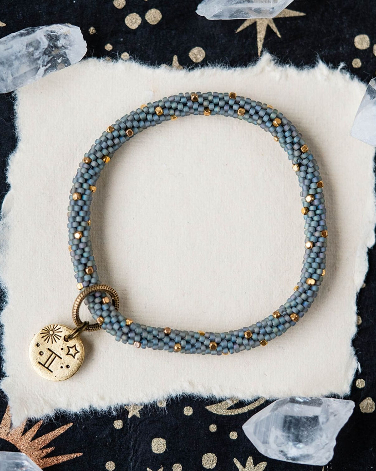 Blue-grey beaded bracelet with gold accents and Gemini zodiac charm on starry background