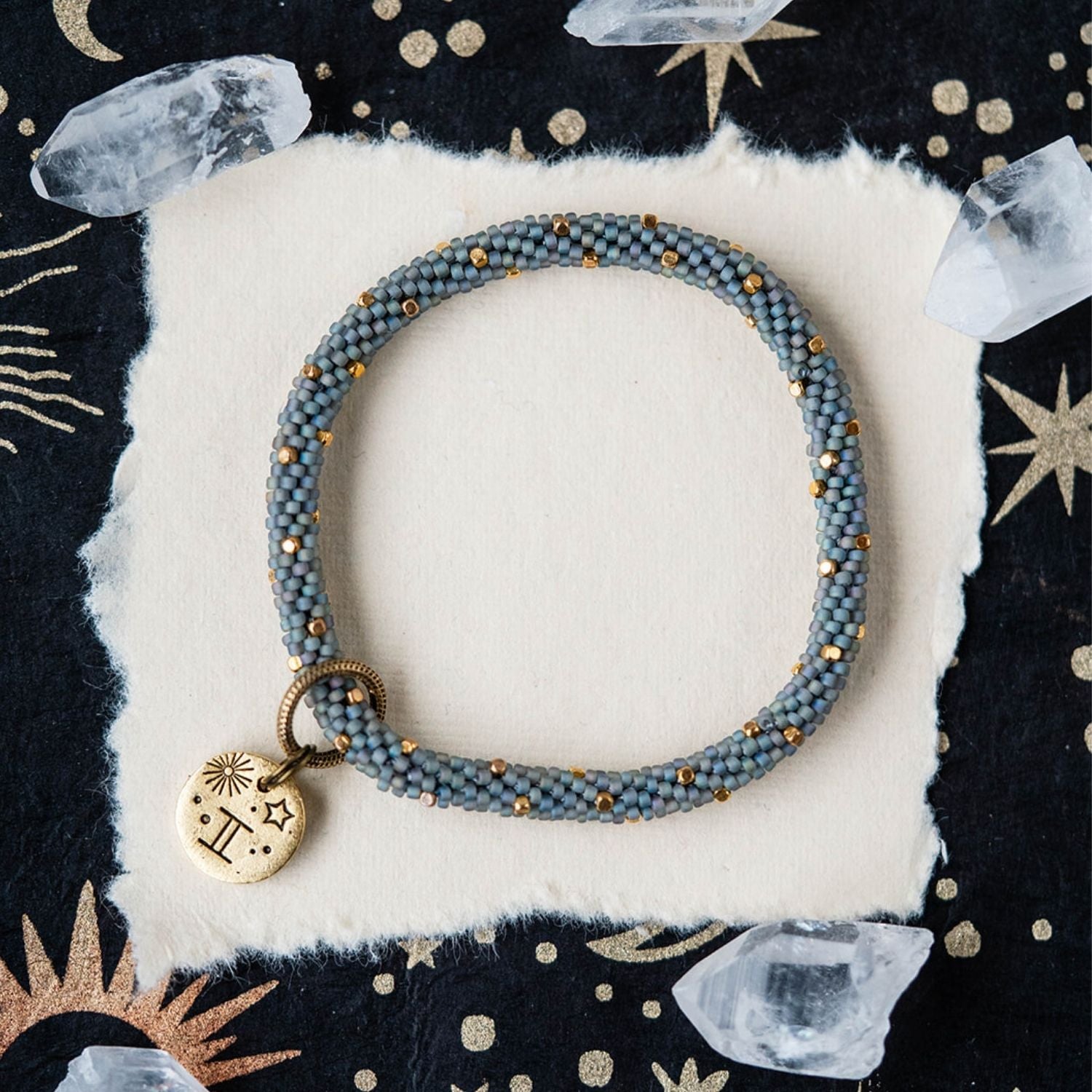 Blue-grey beaded bracelet with gold accents and Gemini zodiac charm on starry background