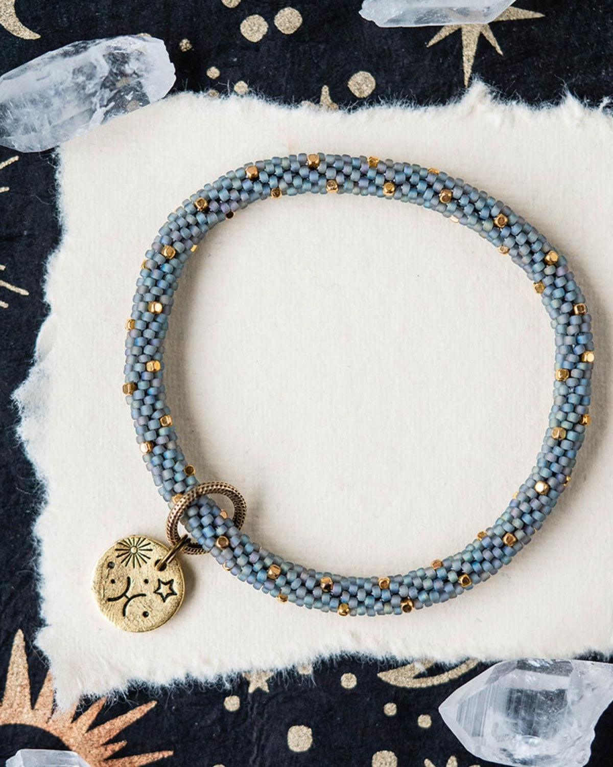 Blue-grey beaded bracelet with gold spacers and Pisces zodiac charm on starry background
