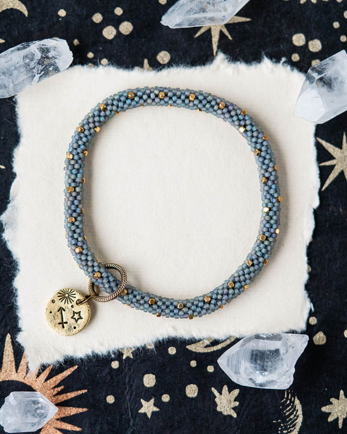 Blue-grey beaded bracelet with gold accents and Sagittarius zodiac charm on celestial background