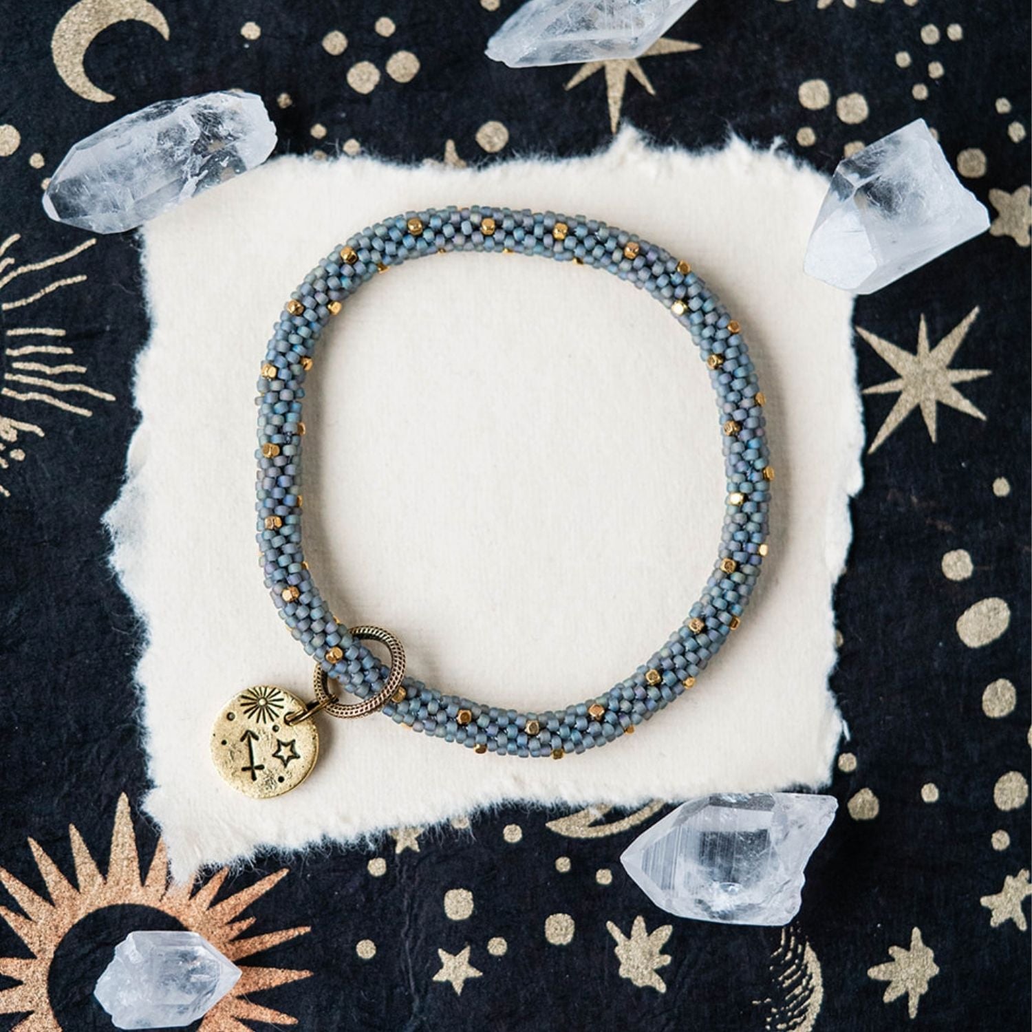 Blue-grey beaded bracelet with gold accents and Sagittarius zodiac charm on celestial background