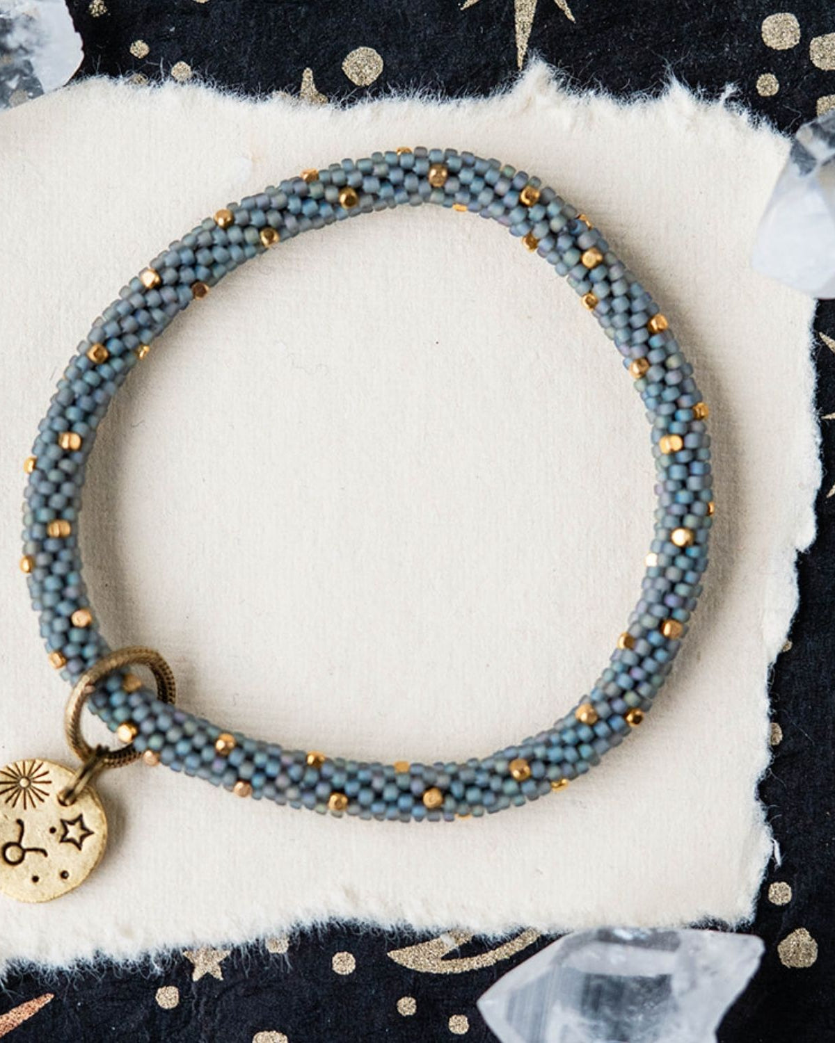 Blue-grey beaded bracelet with gold spacers and Taurus zodiac charm on starry background