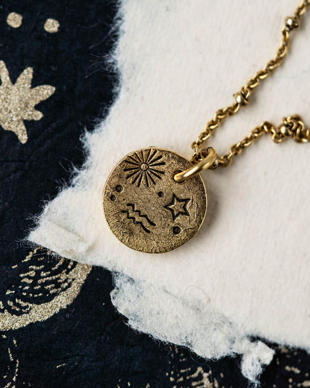Gold Aquarius zodiac pendant necklace with celestial symbols on chain against starry fabric background