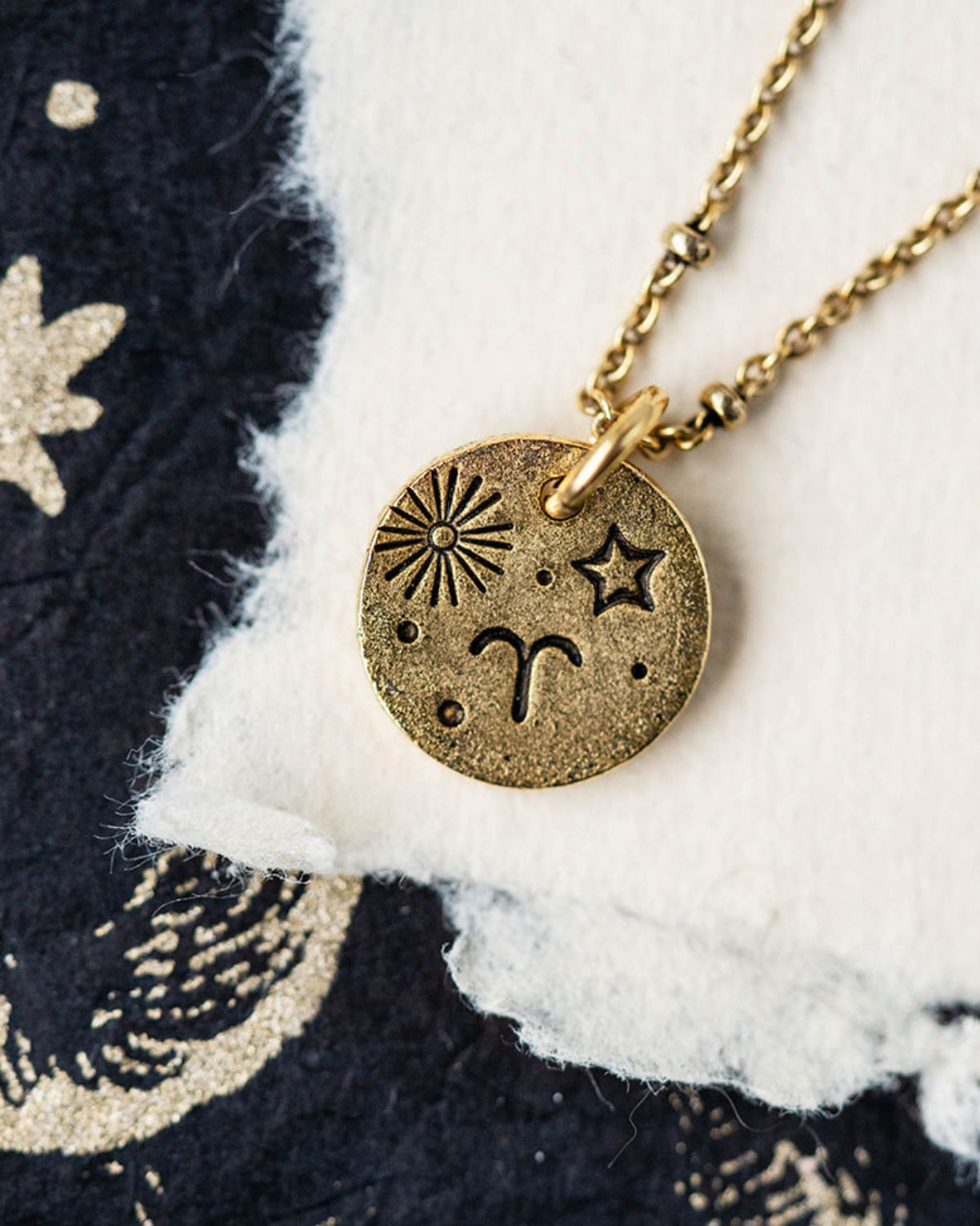 Gold Aries zodiac pendant necklace with ram horn and sun symbols on celestial fabric background