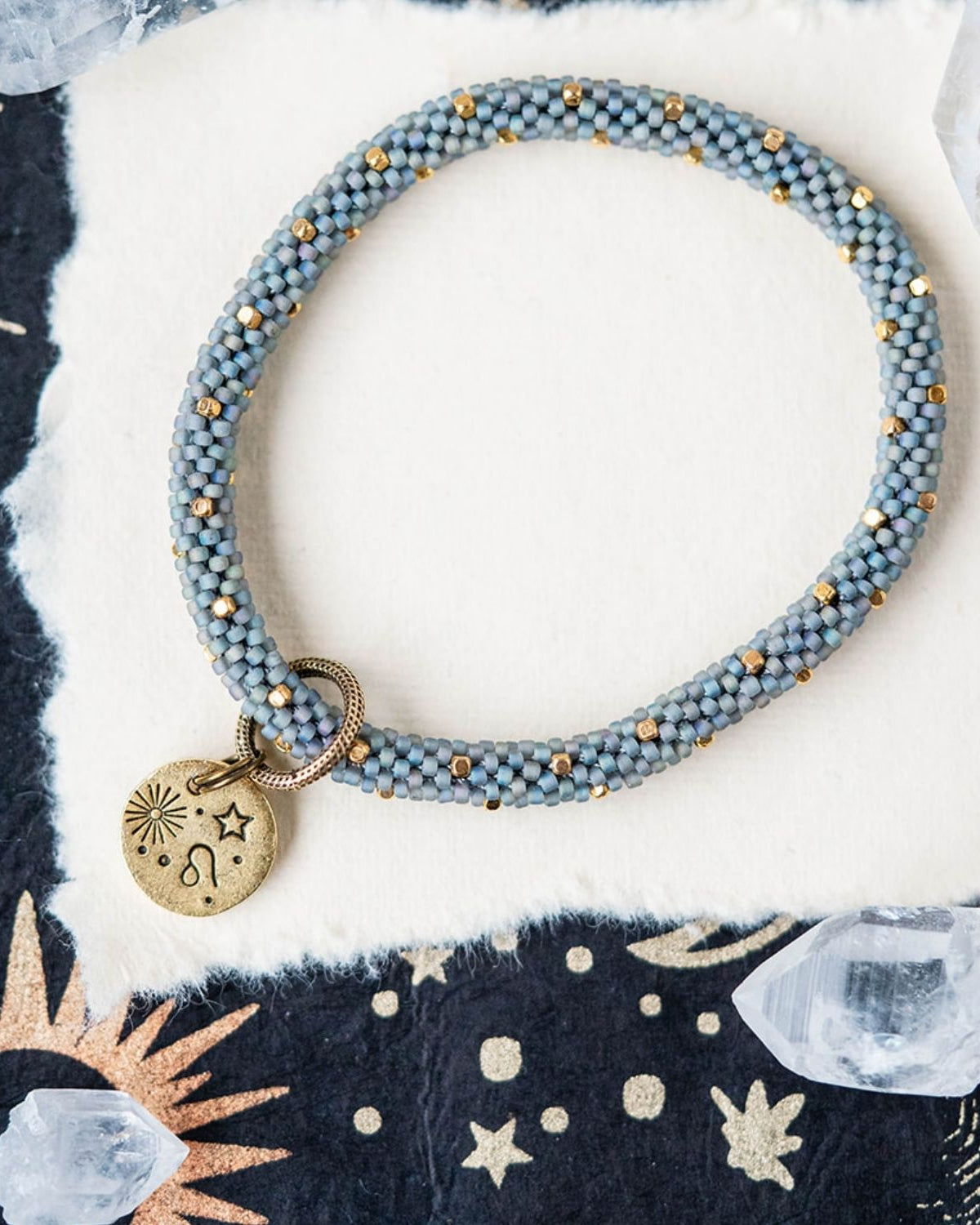 Blue-grey beaded bracelet with gold spacers and Leo zodiac charm on starry background