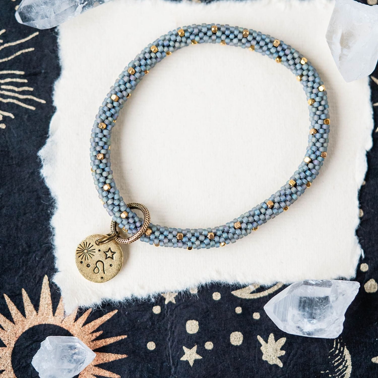Blue-grey beaded bracelet with gold spacers and Leo zodiac charm on starry background