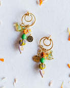 A pair of handmade gold hoop earrings with dangling beaded charms in green, orange, yellow, and white, arranged on a white surface with scattered orange and white flower petals.