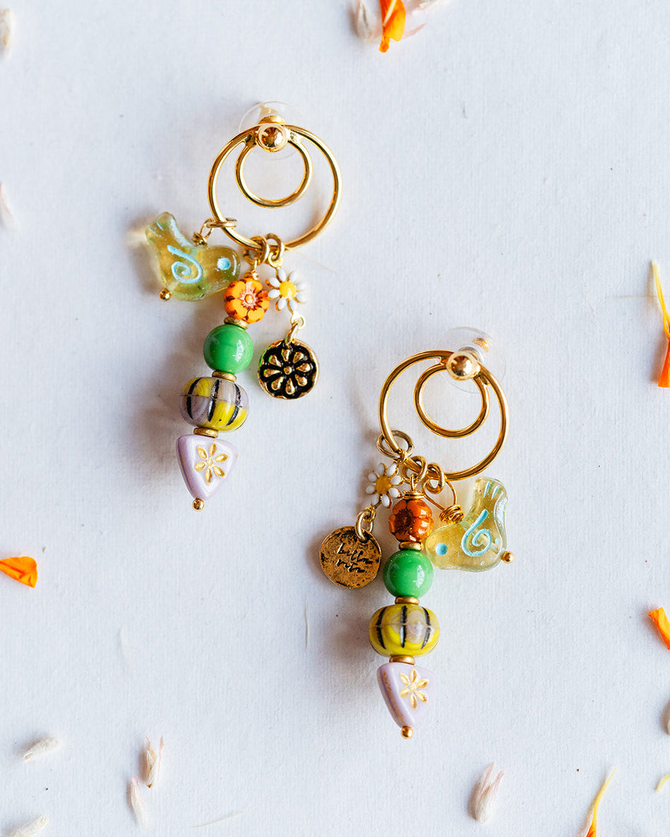 A pair of handmade gold hoop earrings with dangling beaded charms in green, orange, yellow, and white, arranged on a white surface with scattered orange and white flower petals.