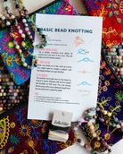 A flat-lay of a 'Basic Bead Knotting' instruction sheet from Bella Vita, surrounded by colorful embroidered pouches and numerous beaded necklaces on a white surface.