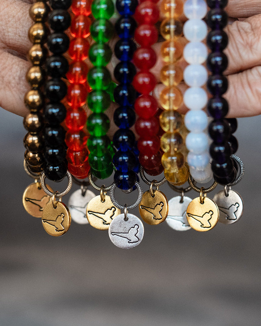 A close-up of two hands holding up a set of colorful beaded bracelets with dangling martial arts kicker charms. The bracelets feature a variety of bead colors including black, gold, red, green, blue, orange, white, and purple, with some charms shown in silver and gold.