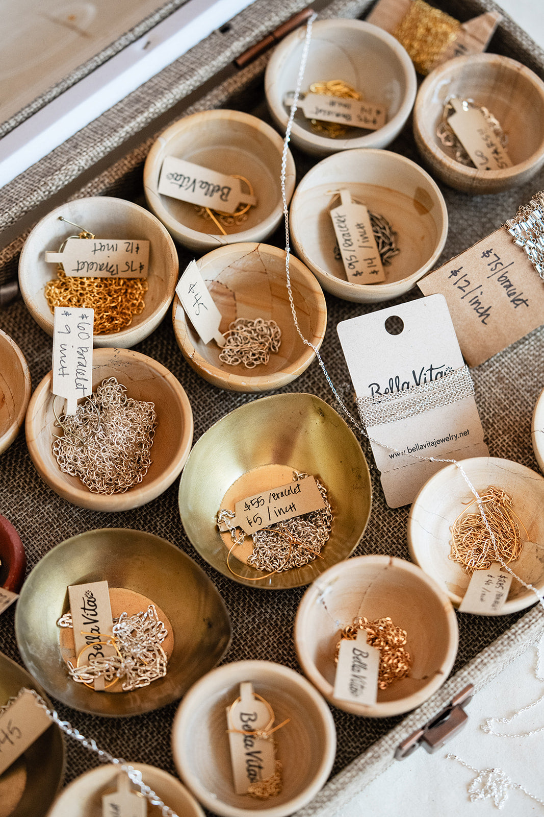 An overhead styled shot of multiple small ceramic bowls in matte and metallic gold and beige tones, each holding a different jewelry piece or material sample with handwritten tags. A white card reading 'BellaVita' is visible among the pieces, and the arrangement sits on a textured neutral surface.