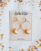 Gold-tone hoop earrings with crescent moon charms, displayed on a Bella Vita jewelry card with dried flower petals on a light surface.