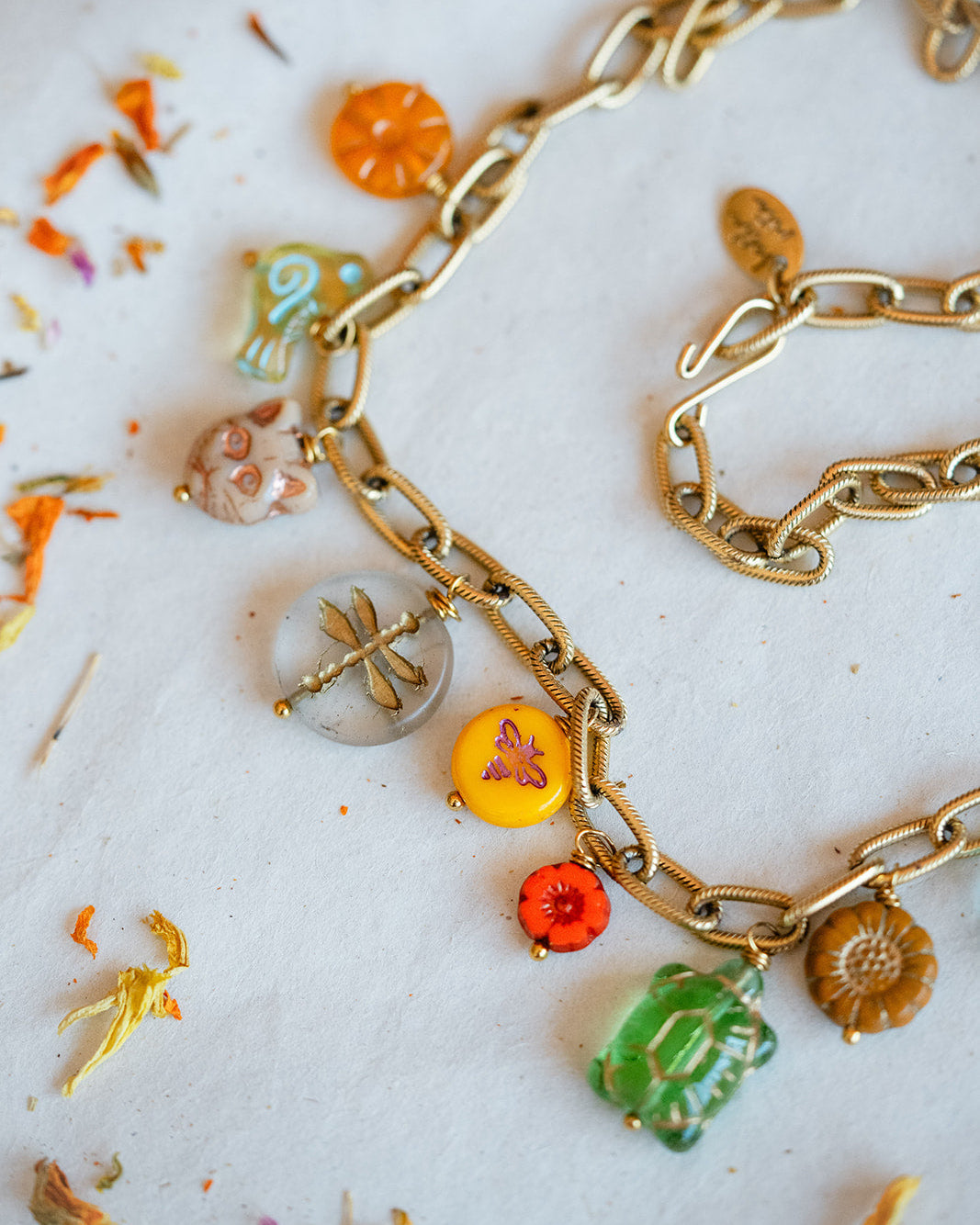 A gold chain necklace with multiple colorful, nature-themed charms including a dragonfly, turtle, butterfly, sunflower, and leaf, displayed on a white surface with scattered dried flower petals.