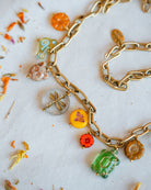 A gold chain necklace with multiple colorful, nature-themed charms including a dragonfly, turtle, butterfly, sunflower, and leaf, displayed on a white surface with scattered dried flower petals.